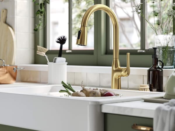 Modern kitchen sink with a gold faucet, green cabinets, cleaning brushes, soap dispenser, and fresh vegetables on the counter.