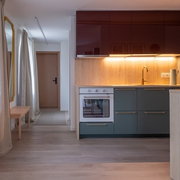 Modern kitchen with a sleek and stylish design featuring dark green lower cabinets with brass handles and glossy burgundy upper cabinets. The backsplash and countertop area is lit by warm under-cabinet lighting, highlighting the natural wood textures. A built-in white oven is integrated into the cabinetry, and a gold faucet adds an elegant touch. To the left, a hallway leads to a light wooden entrance door, with a long mirror, beige curtains, and a small wooden bench placed along the wall. The overall space is minimal yet warm, with a mix of rich colors and natural tones.