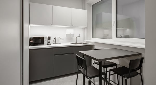 Modern kitchenette with sleek grey cabinets, built-in sink, microwave, coffee machine, kettle, and a black dining table with four chairs.