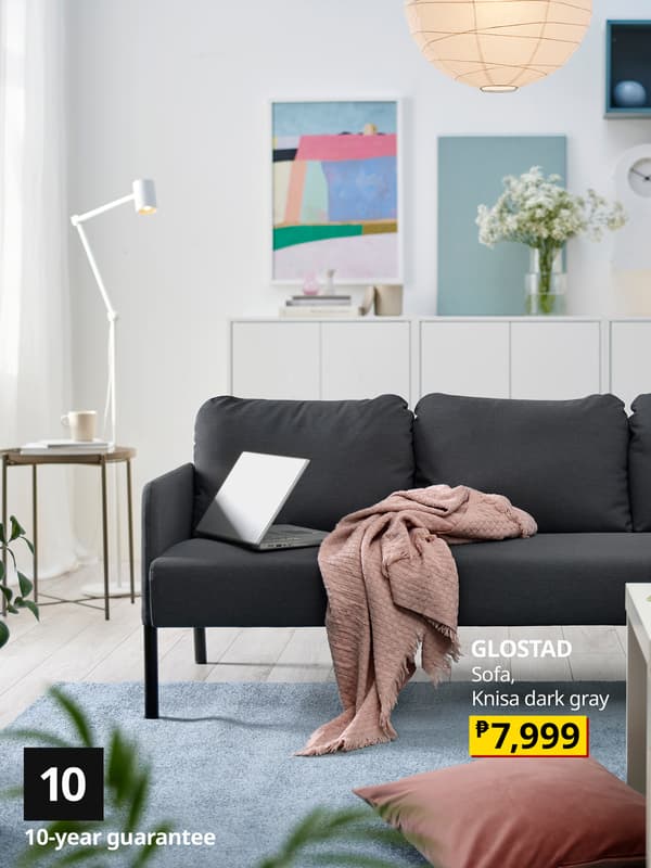 Modern living room featuring GLOSTAD, a dark gray sofa with a pink throw blanket, a nearby lamp, and decorative artwork. Price tag displayed with P7,999.