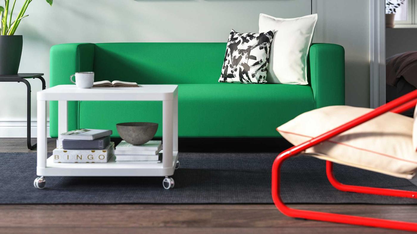 Modern living room with a vibrant green KLIPPAN sofa, styled with black-and-white and cream cushions. A white TINGBY coffee table on castors holds books, a bowl, and a coffee cup, set on a dark rug. A red-framed lounge chair with cream cushions adds a pop of contrast.