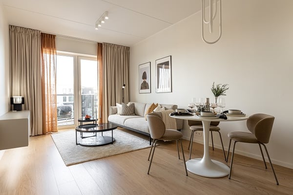 Modern living room with beige sofa, round dining table, and large windows with curtains.