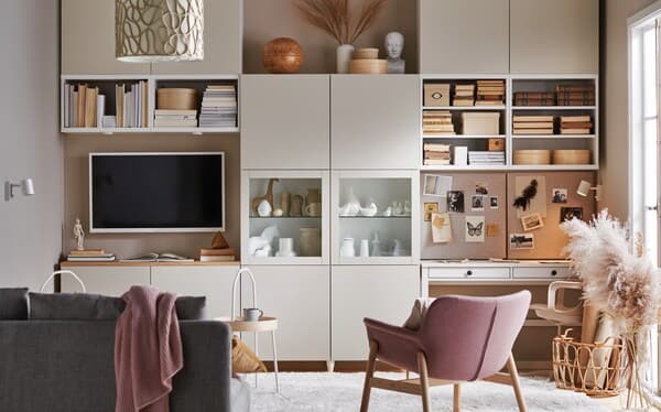 Modern living room with custom storage cabinets, open shelving, and an integrated workspace. The room features a television, decorative accessories, a pink accent chair, a grey sofa, and warm neutral tones.