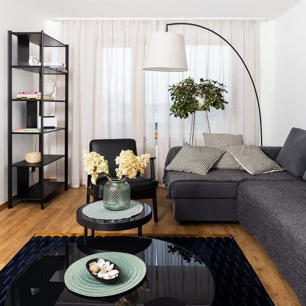 Modern living room with grey sofa and black shelving – Stylish living room with sectional grey sofa, black armchair, decorative coffee tables, green plant, and floor lamp against sheer curtains.
