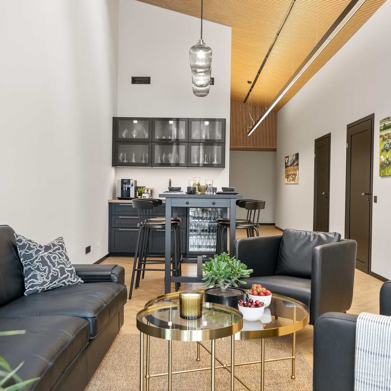 Modern lounge area with black leather chairs and a round coffee table holding plants and bowls of fruit, leading to a small kitchen area with dark cabinets, high stools, and pendant lights under a slatted wooden ceiling.