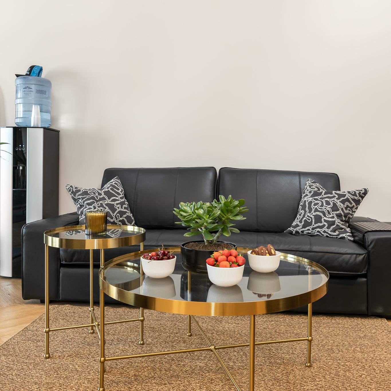 Modern lounge area with black leather seating arranged around gold-framed glass coffee tables holding a decorative plant and bowls of snacks.