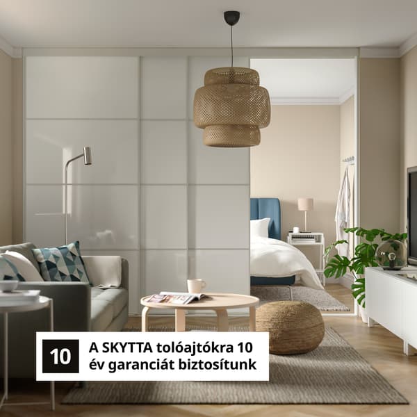 Modern nappali, amely a hálószobával kapcsolódik, fehér SKYTTA tolóajtókkal elválasztva, világos belső tér kanapéval, dohányzóasztallal, függesztett rattan lámpával és 10 éves garanciáról szóló szöveggel.