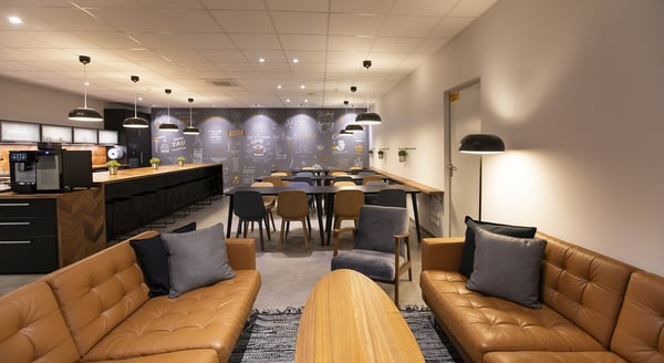 Modern office break room with leather sofas, wood coffee table, mixed dining chairs, long bar counter with black stools, pendant lighting, coffee station, and chalkboard-style wall art.