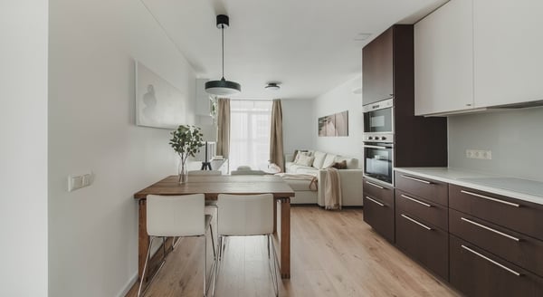 Modern open-concept kitchen and living room with a warm, minimalist design. The kitchen features sleek dark wood lower cabinets and integrated appliances, contrasted by white upper cabinets. A wooden dining table with white chairs separates the kitchen from the living area. Soft brown curtains, a neutral-toned sectional sofa, and soft lighting create a cozy ambiance.