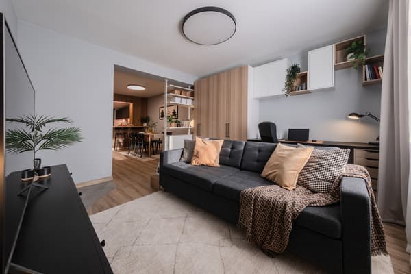 Modern open-plan living room with a grey sofa, light wood flooring and built-in shelving, flowing into a kitchen and dining area, with a workspace featuring a desk and chair in the background.