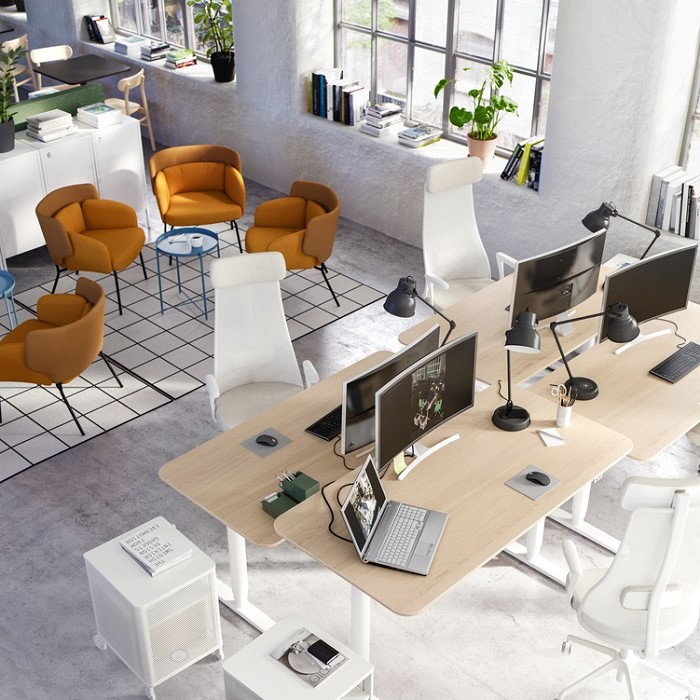 Modern open-plan office with shared workstations, ergonomic white chairs, dual monitors, and a cozy lounge area with mustard yellow armchairs by large industrial windows.