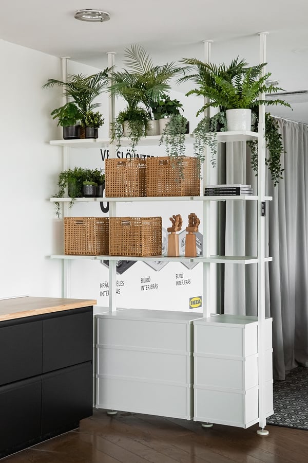 Modern open shelving unit with white frame and drawers, styled with lush green potted plants, woven rattan baskets, wooden decorative figures, and neatly stacked books in a bright IKEA showroom space.
