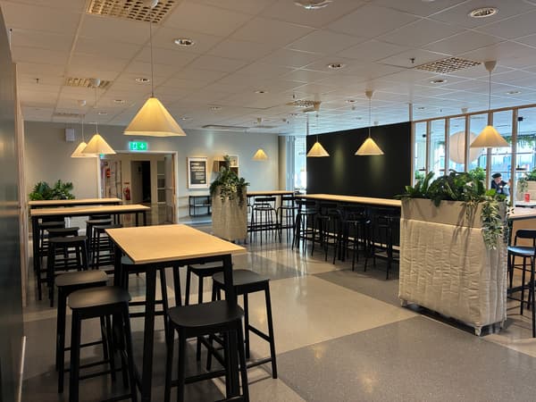 Modern personalmatsal med höga bord, svarta barpallar, växtinslag och hänglampor i en öppen och luftig miljö.
