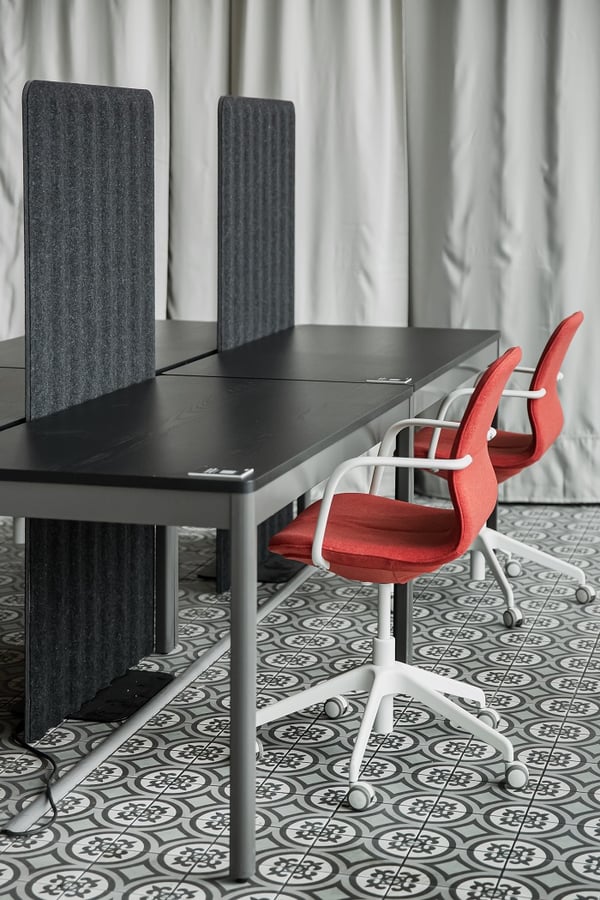 Modern shared workspace with black desks, gray felt privacy panels, red swivel office chairs with white frames, and decorative patterned tile flooring in a clean, minimalist environment.