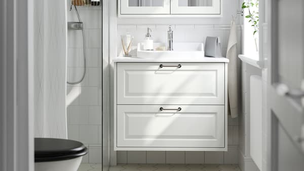 Modern TÄNNFORSEN bathroom with white tiles, white bathroom furniture with washbasin and toiletries, shower with glass door, mirror cabinet and natural light through a window.