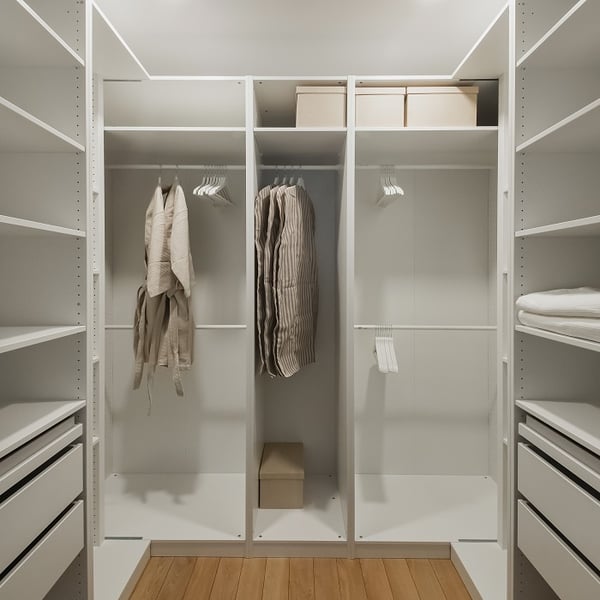 Modern walk-in closet with white open shelving and hanging space. Several beige clothing items and striped garments hang neatly, alongside white hangers. Storage boxes are placed on the top shelves, and folded linens rest on the right side shelves. The wooden floor adds warmth to the minimalist design.