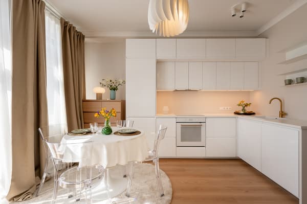 Modern white kitchen with a round dining table set for four, transparent chairs, wooden flooring, and beige curtains.