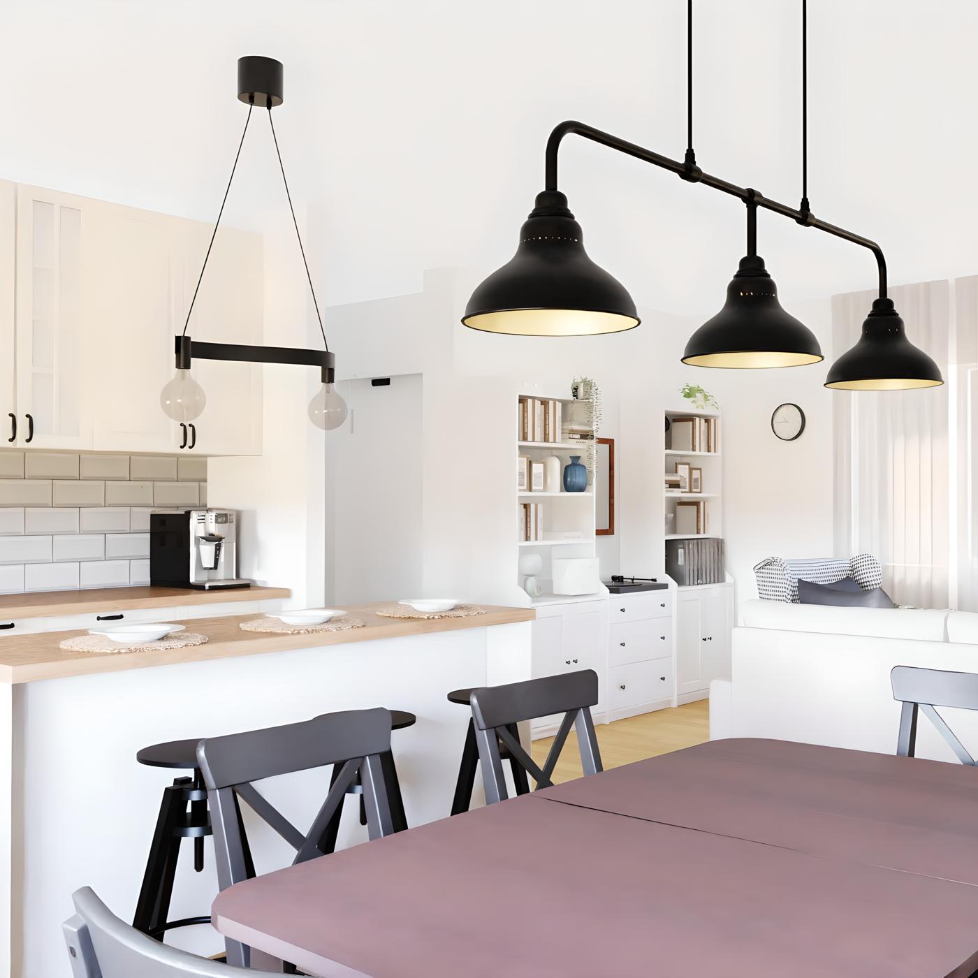 Moderna kuhinja i blagovaonica s drvenim stolom, crnim stolicama i visećim svjetiljkama; u pozadini dnevni boravak s bijelim namještajem, policama za knjige i zidnim satom.