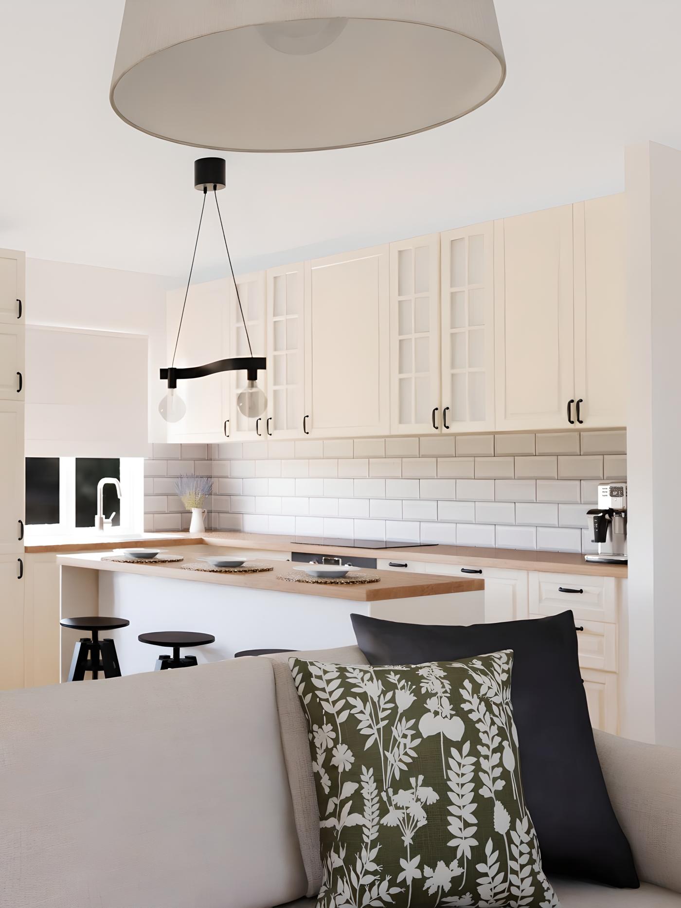 Moderna kuhinja s bijelim ormarićima, drvenim pločama i bijelim pločicama; u prvom planu bež kauč s jastucima, iznad kuhinjskog otoka dvije viseće žarulje na crnoj šipki.