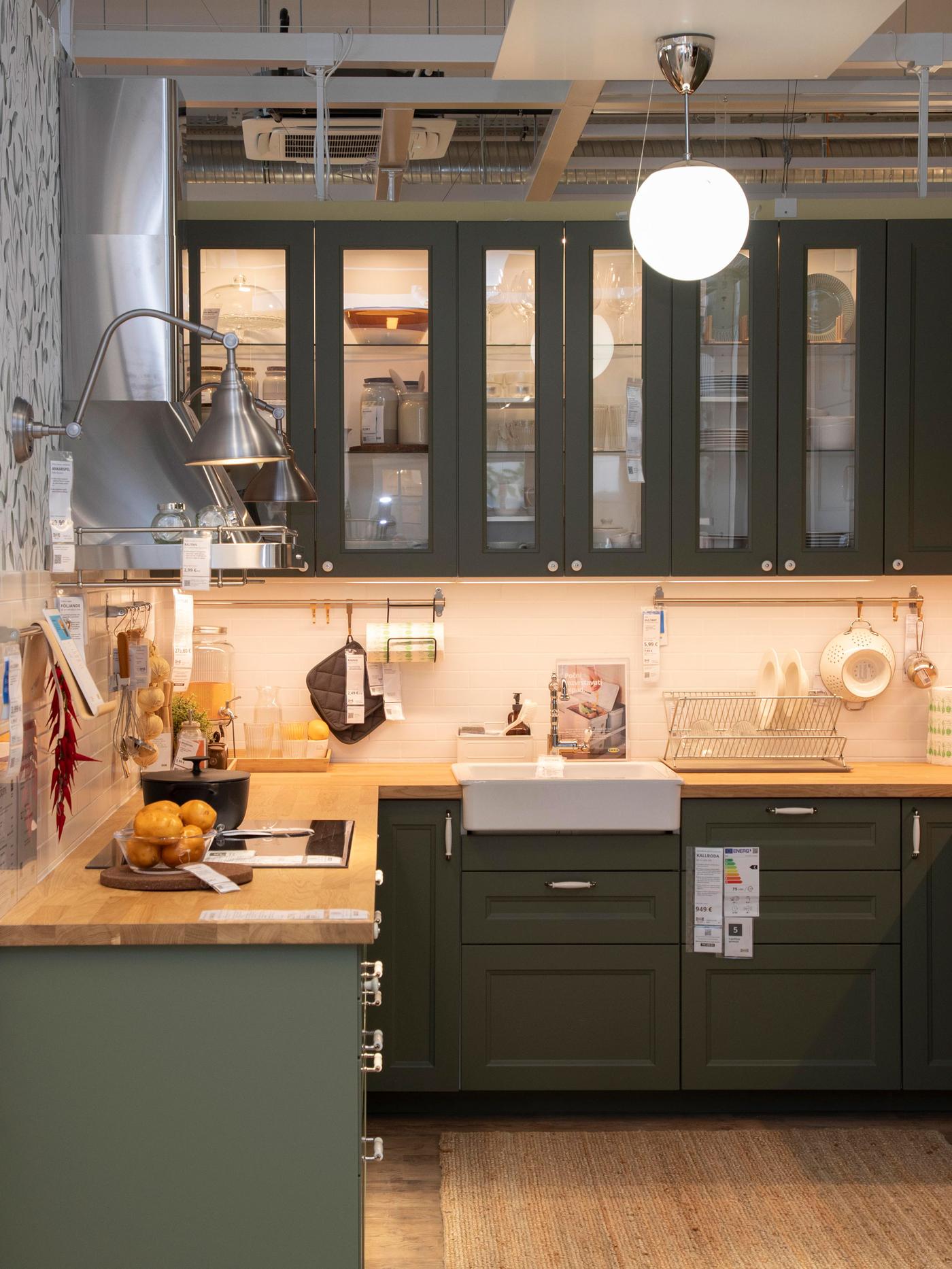 Moderna kuhinja s tamnozelenim ormarićima, drvenim radnim pločama i bijelim sudoperom; na ploči voće, začini i pribor, iznad ormarića stakleni dio s posuđem, topla rasvjeta i okrugla lampa na stropu.