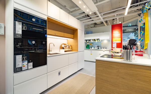 Moderne IKEA-Küchenausstellung mit weißer Küchenzeile und Kochinsel mit Holzoptik. Im Vordergrund Küchengeräte und Utensilien, im Hintergrund weitere Küchenbereiche und Deko mit schwedischen Flaggen.

