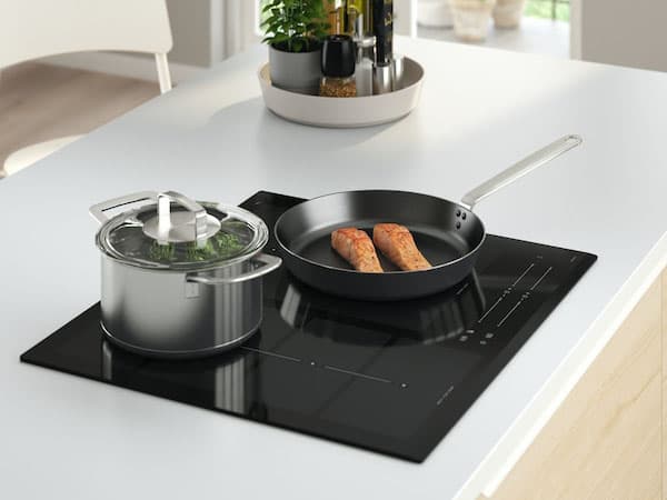 Moderne Küchenarbeitsplatte mit Induktionskochfeld; darauf ein Topf mit Glasdeckel, gefüllt mit grünem Gemüse, und eine Pfanne mit zwei Lachsfilets. Im Hintergrund steht ein runder Servierteller mit Flaschen und Küchenutensilien.
