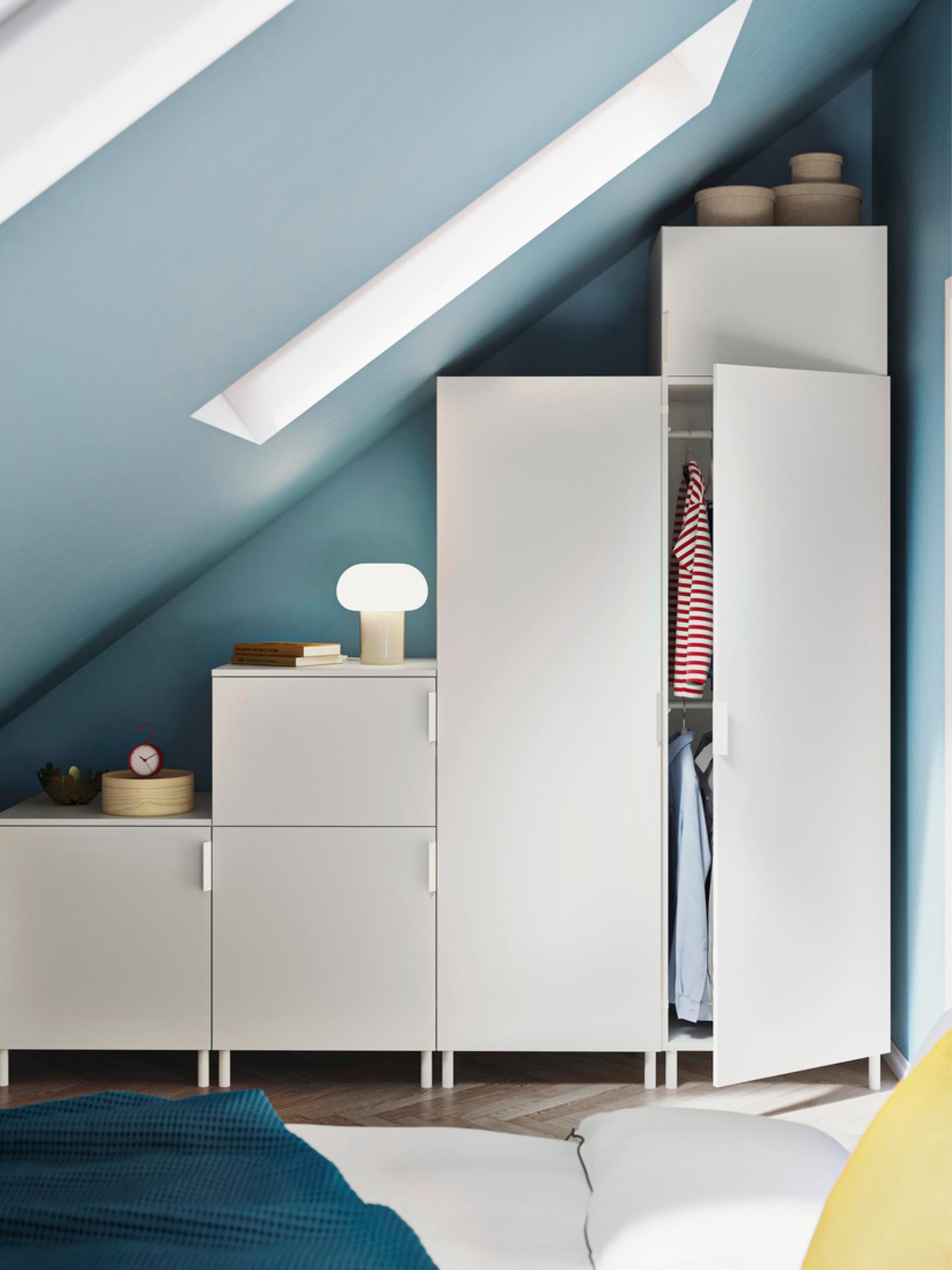 Moderne slaapkamer met schuin plafond en blauwe muren, met witte modulaire kledingkasten, open kast met hangende kleding en een bed met wit beddengoed, blauwe deken en geel kussen.