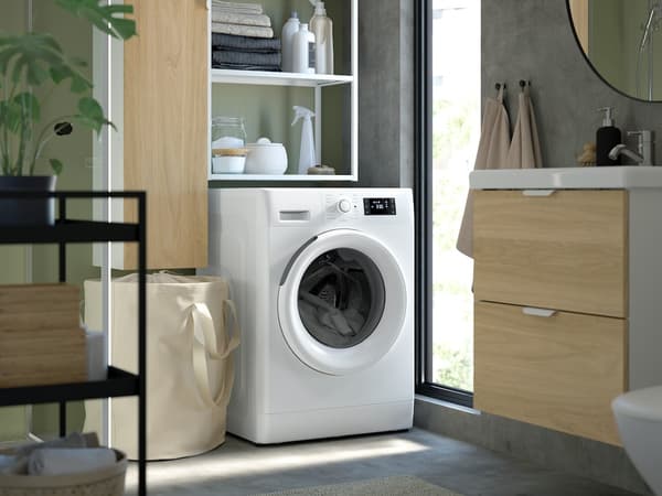 Moderne Waschecke mit weißer Frontlader-Waschmaschine zwischen Holzschränken; daneben ein Wäschekorb, darüber offene Regale mit Haushaltsartikeln.