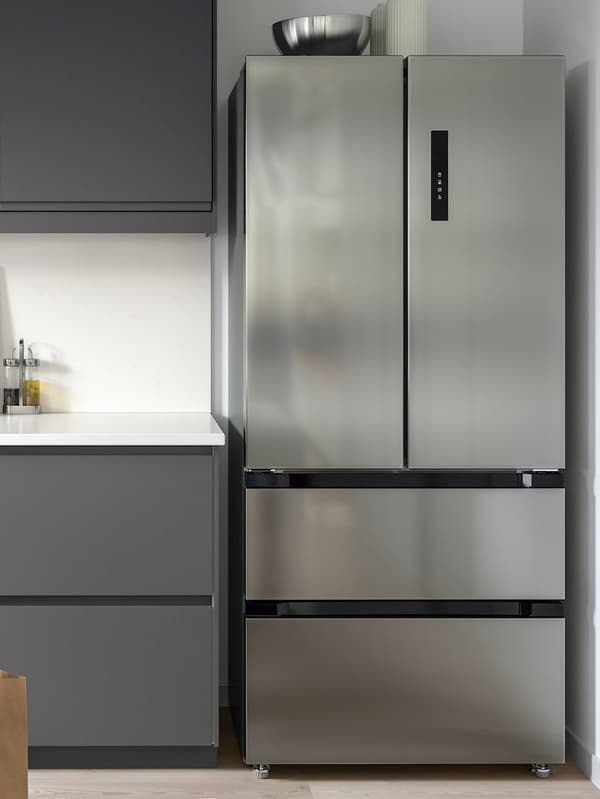 Moderner Kühlschrank aus Edelstahl mit zwei Türen oben und zwei Schubladen unten, eingebaut in eine Küche neben einem dunkel gefärbten Schrank und einer Arbeitsplatte.