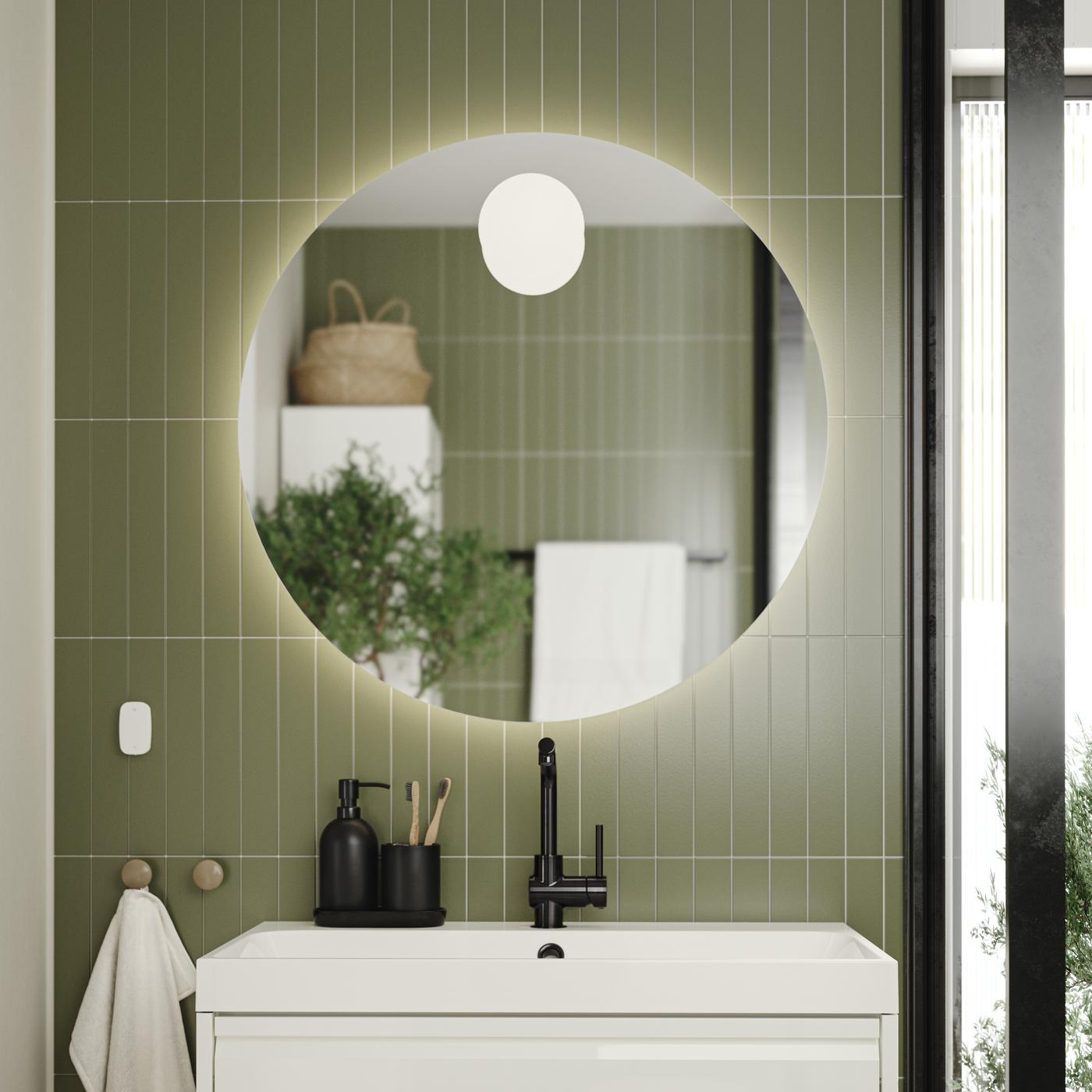 Modernes Badezimmer mit rundem, beleuchtetem FAXÄLVEN Spiegel an grüner Fliesenwand, weißem Waschbecken mit schwarzem Wasserhahn, Handtuch links, Fenster oder Glastür mit Blick ins Grüne rechts und Badaccessoires auf dem Waschbecken.