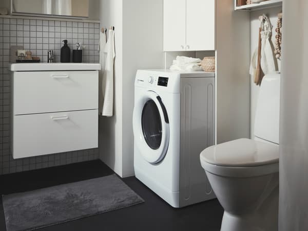 Modernes Badezimmer mit Waschmaschine, weißem WC und Waschtisch mit zwei Schubladen; auf der Ablage stehen Pflegeprodukte, davor liegt ein dunkler Teppich.