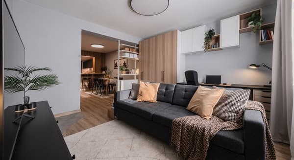 Moderni atviro plano svetainė su pilka sofa, šviesiomis medinėmis grindimis ir įmontuotomis lentynomis, pereinanti į virtuvę ir valgomojo zoną. Už sofos matosi darbo vieta su stalu ir kėde.