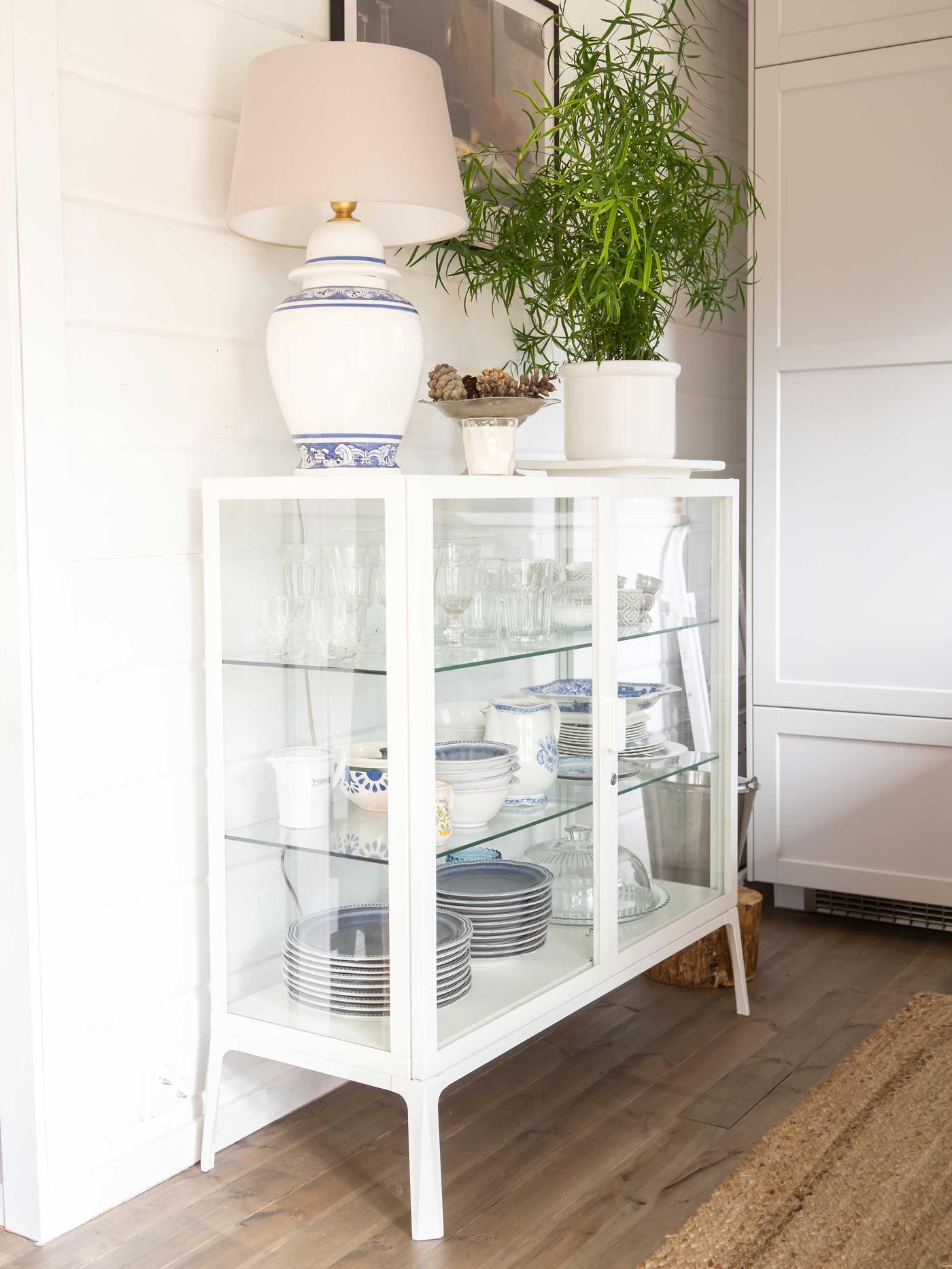 Modernin mökin keittiössä on valkoinen vitriinikaappi, jonka päällä on viherkasvi ja pöytävalaisin.