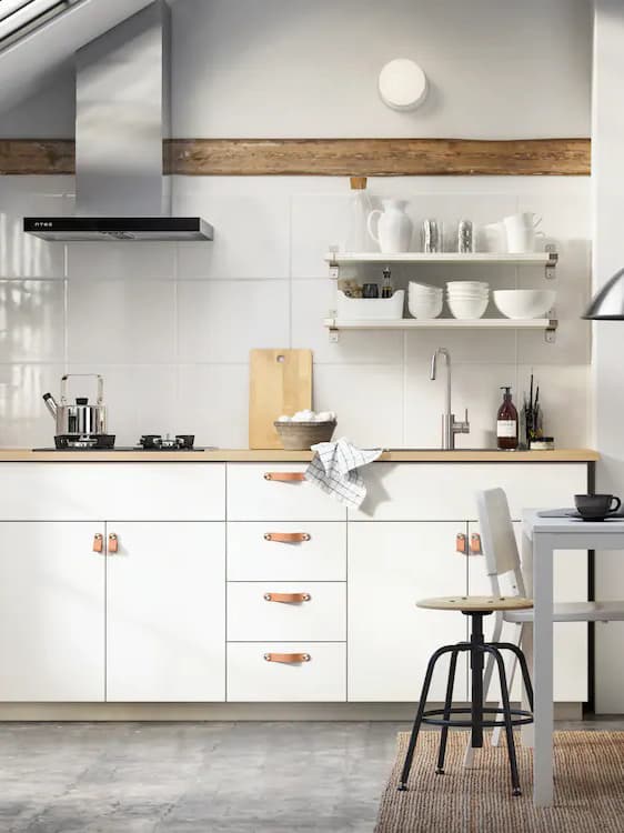 Modernt kök med vita skåp, läderhandtag och öppna hyllor; spis, diskho och köksredskap syns, med ljusinsläpp från vänster.