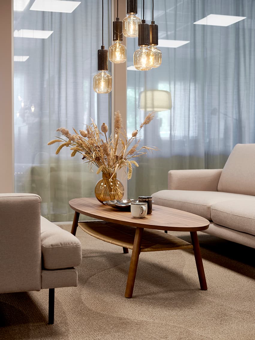 Modernt vardagsrum med beige soffa, träbord med vas och böcker, hängande lampor och stora fönster med skira gardiner.