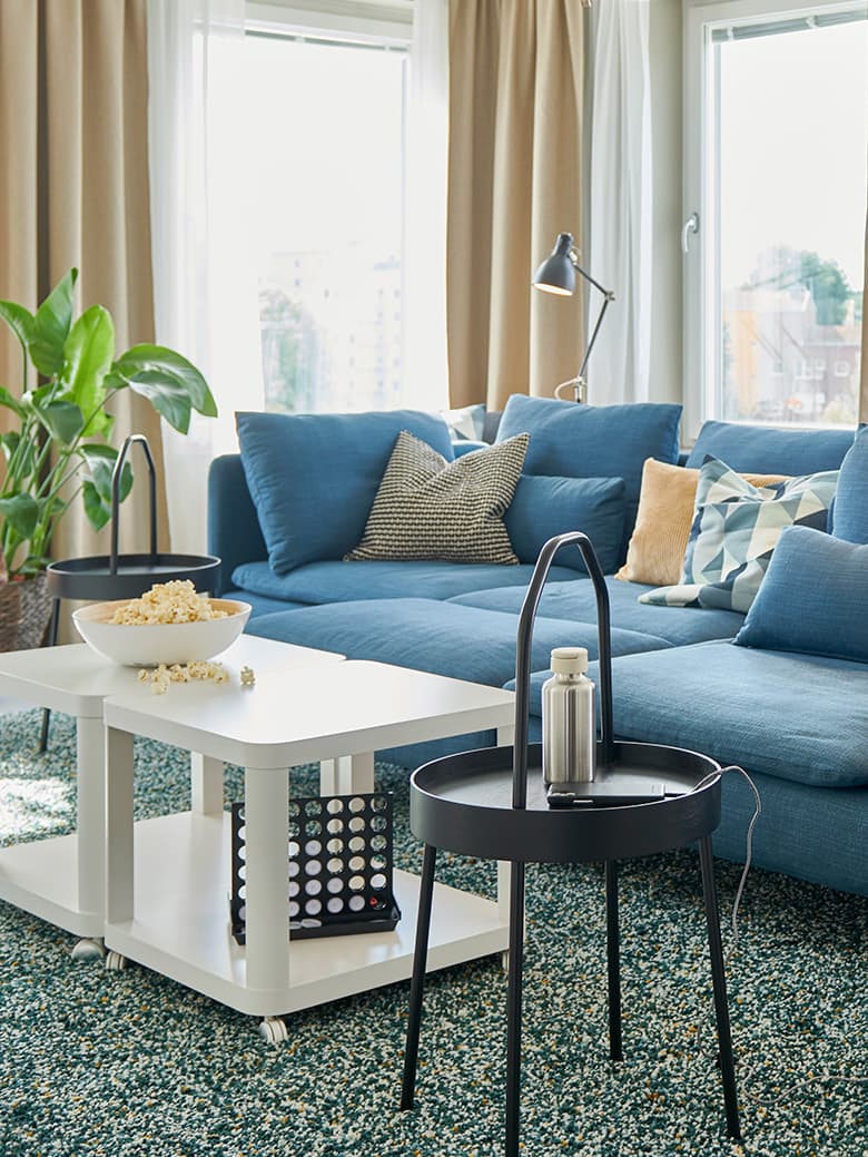 Modernt vardagsrum med blå soffa, vitt soffbord, växter, stora fönster med beige gardiner och mönstrad matta.