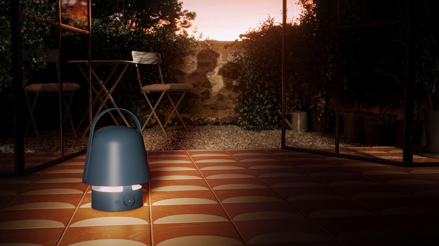 Modrá lampa s Bluetooth reproduktorem VAPPEBY stojí na dlažbě u vstupu do zahrady obehnané zdí; na zdi je stín páru.