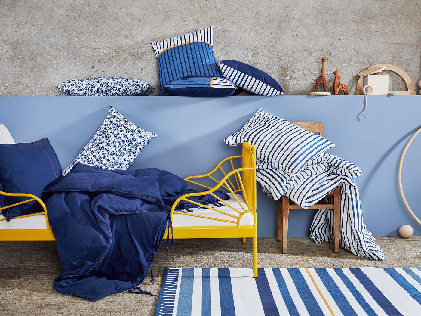 Modro-biela kolekcia textílií SÅNGLÄRKA vedľa detskej postele obsahuje vankúše a posteľné obliečky s pásikavým a kvetinovým vzorom.