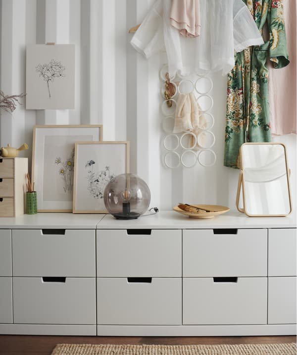 Module de rangement bas blanc sur lequel sont posés des dessins, une lampe et un miroir et vêtements accrochés en hauteur, sur une paroi en tôle ondulée.