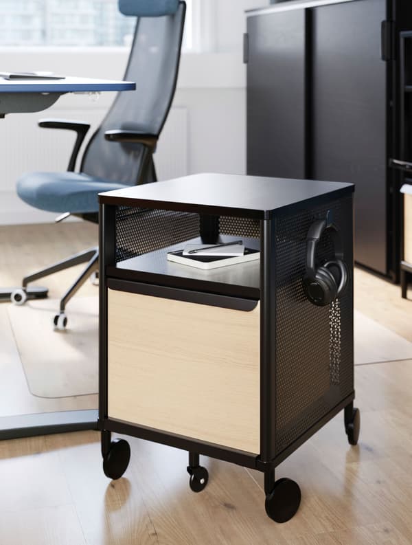 Módulo de almacenaje BEKANT negro con ruedas en negro con un cuaderno y unos auriculares, delante de una silla de trabajo JÄRVFJÄLLET azul.