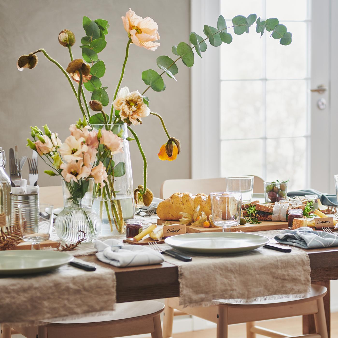 MÖRBYLÅNGA tafel in eiken fineer gedekt voor het eten met mat groen FÄRGKLAR eetgerei en twee bloemschikkingen in glazen vazen.