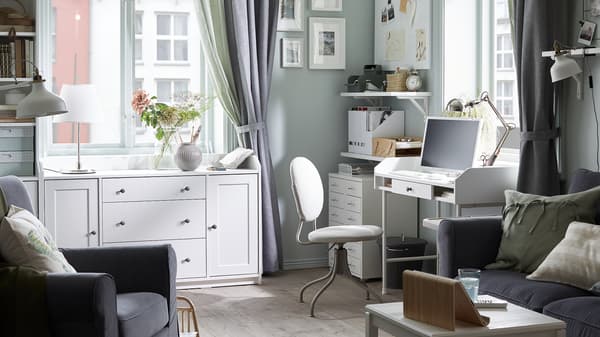 Mörkgrå soffa, vitt skrivbord med en dator, beige kontorsstol, vita hyllor med diverse papper, vita skåp.
