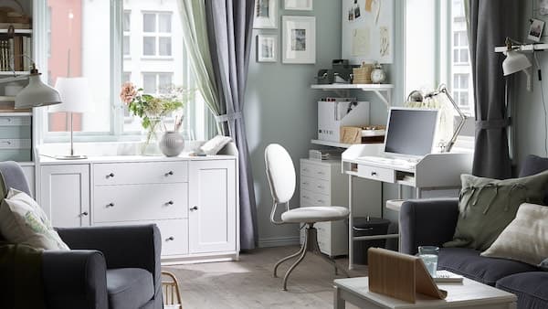 Mörkgrå soffa, vitt skrivbord med en dator, beige kontorsstol, vita hyllor med diverse papper, vita skåp.