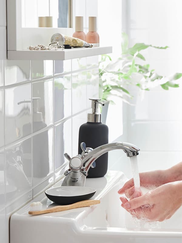 Mosdó egy EKOLN sötétszürke tállal és EKOLN szappanadagolóval, és valaki a kezét mossa a RUNSKÄR csapból folyó vízben.