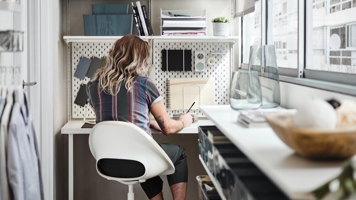 Moški na bež/belem vrtljivem stolu za belo mizo z belim sistemom za shranjevanje zgoraj, na zaprtem balkonu.