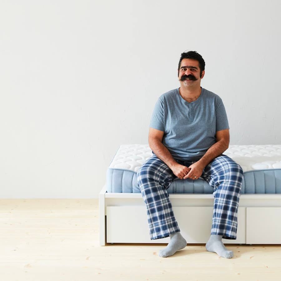Mosolygós, bajuszos, pizsamás férfi ül egy matracon.