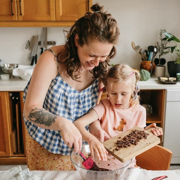 Mother and daughter at the kitchen table. The adult helps the child pour chopped chocolate from a wooden board into a bowl.