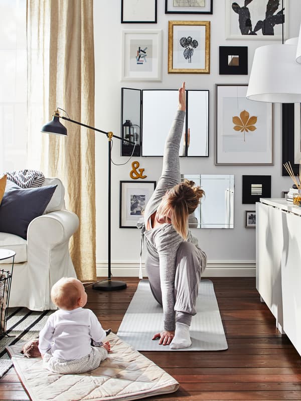 Mother doing yoga on a mat in a living room with a baby sitting on a mat watching her with sofa, sideboard and pictures.