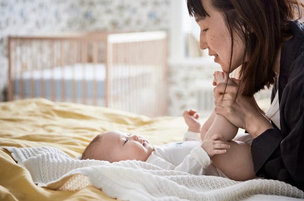 Mother holding her baby by the toes and smiling as the baby lies on yellow bedsheets with a cot in the background.