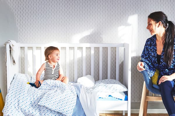 Mother sitting on a wooden chair and baby sitting in white open-sided crib with blue textiles both looking at each other.